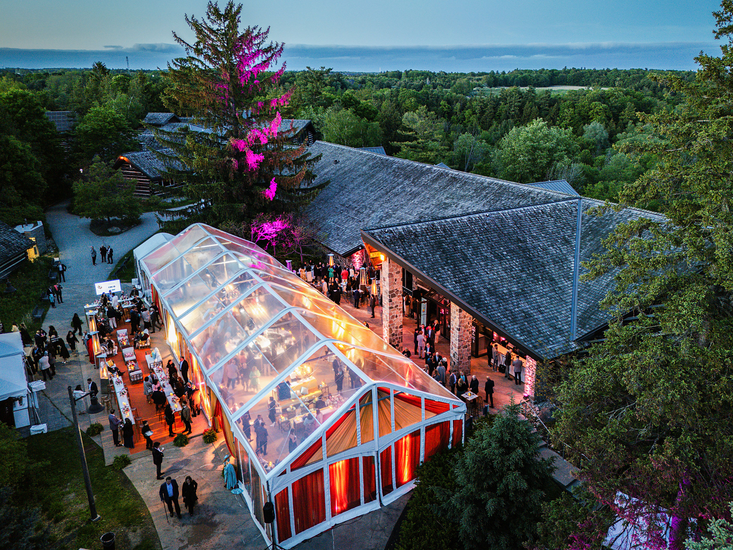 Outdoor evening event with a large transparent tent lit in warm colors, surrounded by people, vendor tables, and the rustic stone McMichael building, with trees and pink accent lighting in the background.